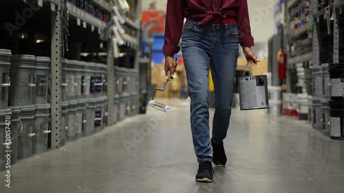 Woman shown from waist down, carrying can of paint and roller walking down paint aisle in hardware store