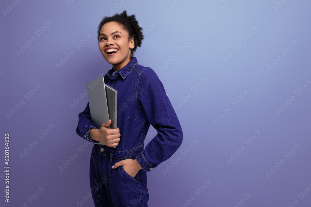 young joyful hispanic business woman with fluffy hair dressed in a blue denim suit works in the office and holds a laptop in her hands young joyful hispanic business woman with fluffy hair dressed in a blue denim suit works in the office and holds a laptop in her hands