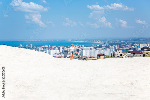 A young woman in a white tank top and denim shorts stands against the backdrop of a resort town on the sea on a sunny day. Magnificent landscape.