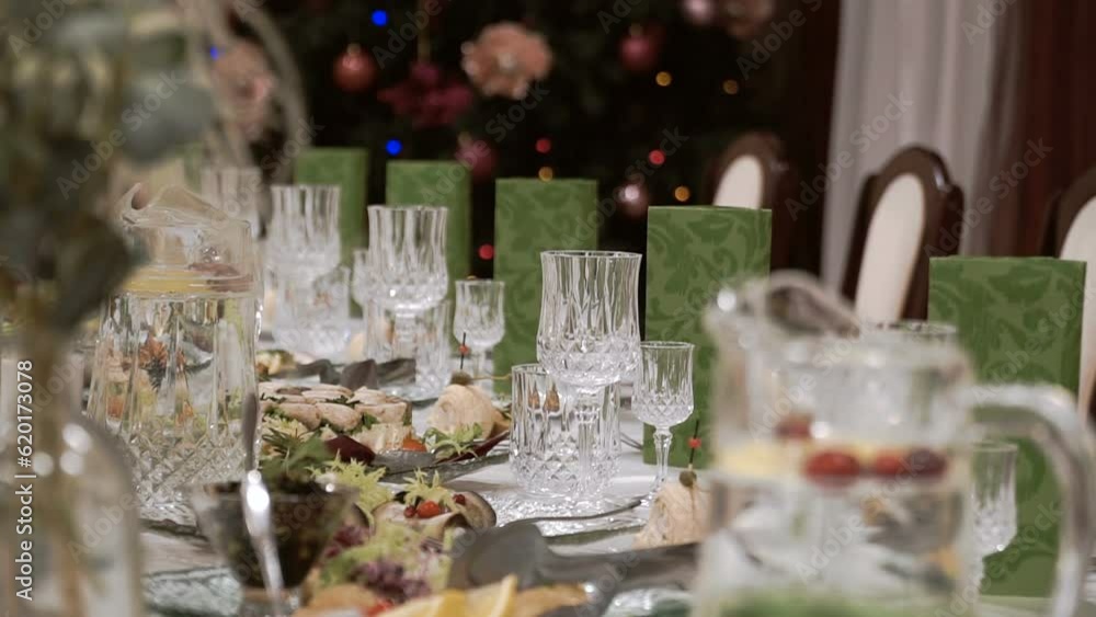 Glass goblets on the banquet table. Table setting with crystal glasses and green napkins. Festive banquet. New Year's lights of Christmas garlands on a tree. Glass goblets on the banquet table. Table setting with crystal glasses and green napkins. Festive banquet. New Year's lights of Christmas garlands on a tree.