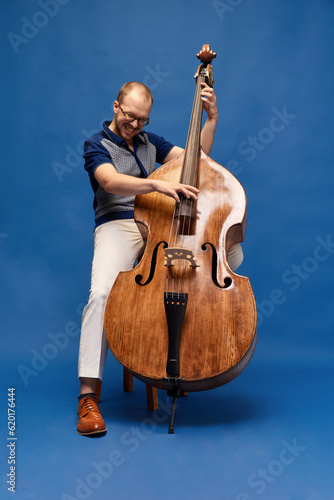 Artistic, emotional man in elegant clothes playing double bass against blue studio background. Classical style. Concept of music, talent, hobby, entertainment, festival, performance, ad