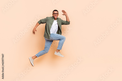 Full length photo of pretty excited man wear khaki shirt running hurrying lumping high empty space isolated beige color background