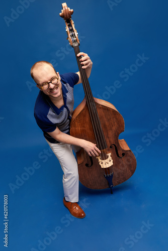 Top view image of artistic young man with moustaches emotionally playing double bass against blue studio background. Concept of music, talent, hobby, entertainment, festival, performance, ad