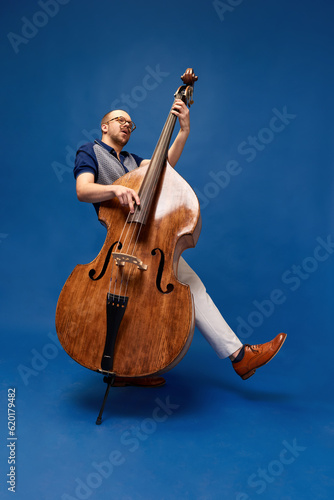 Full-length portrait of young man, musician, emotionally, excessively playing double bass against blue studio background. Concept of music, talent, hobby, entertainment, festival, performance, ad