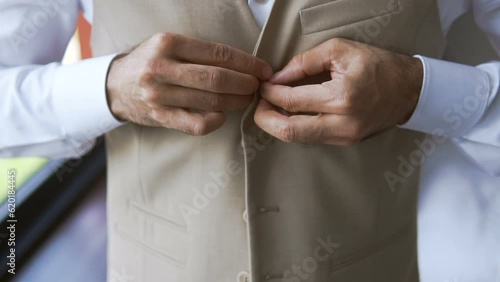 A young businessman puts on a suit before an important meeting. The manager gets dressed near the window in the morning before work. Fingers fasten buttons on a jacket or vest. Beige suit with belt photo