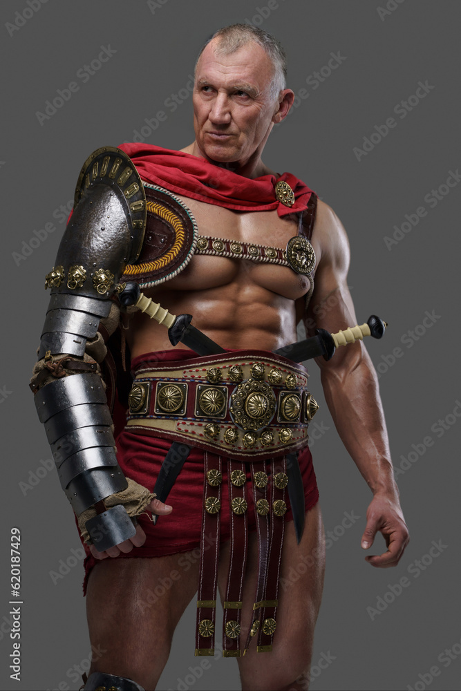 Seasoned and muscular gladiator of mature years with a rough appearance wears armor as he stands confidently against a grey background Seasoned and muscular gladiator of mature years with a rough appearance wears armor as he stands confidently against a grey background