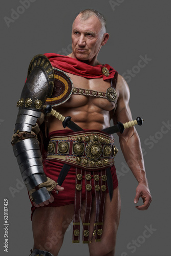 Seasoned and muscular gladiator of mature years with a rough appearance wears armor as he stands confidently against a grey background