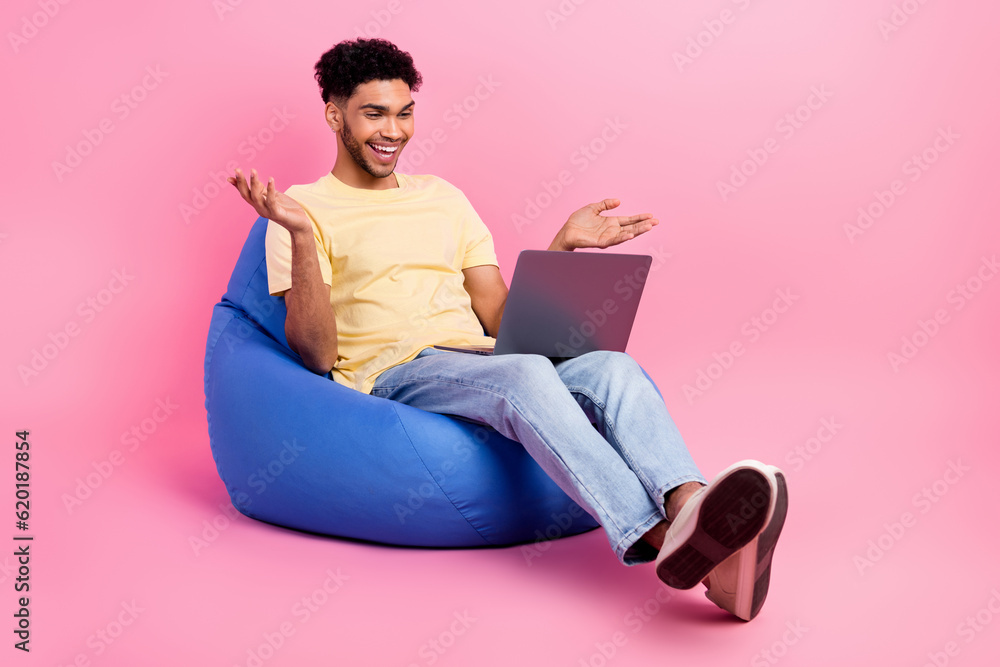 Full length cadre of laughing positive good mood guy sit dorm room with netbook skype call with parents isolated on pink color background Full length cadre of laughing positive good mood guy sit dorm room with netbook skype call with parents isolated on pink color background