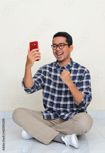 A man sitting cross legged on the floor showing excitement when looking to his mobile phone photo