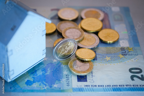 Euro coins resting on banknotes, close up partial view of small wooden house, concept of 'real estate costs' or 'variable mortgages' or 'property market' in europe.