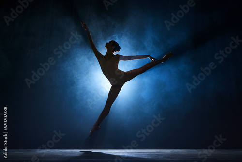Slim, beautiful, graceful ballerina making creative performance on stage against dark blue background with spotlight