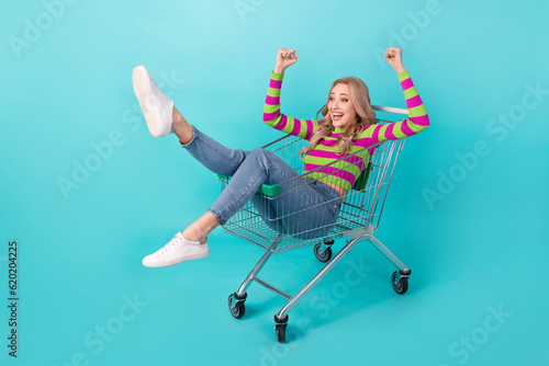 Full length photo of cheerful lucky woman dressed striped sweater driving shopping tray rising fists isolated turquoise color background