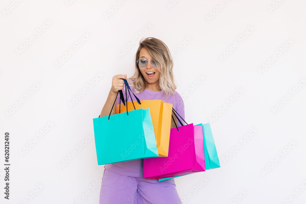 Young attractive amazed caucasian smiling blonde woman in purple outfit and blue stylish sunglasses looks into the color paper bags with purchases isolated on white background. Shopping and fashion