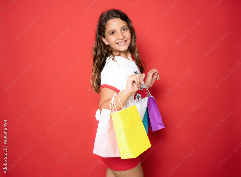 Beautiful brunette little girl holding shopping bags isolated over red background. Beautiful brunette little girl holding shopping bags isolated over red background.
