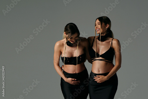 Happy, beautiful pregnant twins sisters in black fashionable and elegant clothes posing together, smiling and touching their bellies. Young women expecting babies.