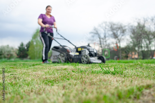 A young woman is mowing a lawn with a lawn mower in his beautiful green floral summer garden. A professional gardener with a lawnmower cares for the grass in the backyard. High quality photo