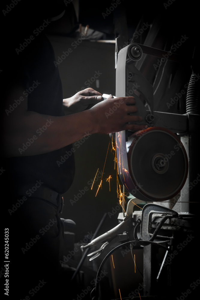 Fototapeta premium Man in a dark forge grinding a knife with a belt grinder and sparks flying downwards