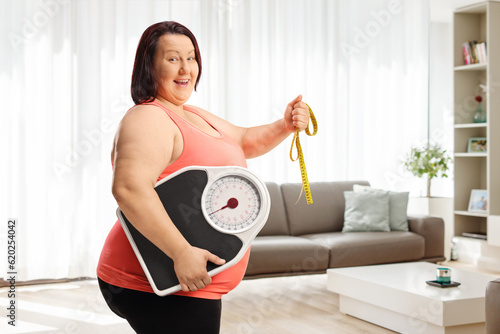 Smiling overweight woman holding a weight scale and a measuring tape inside a livng room photo
