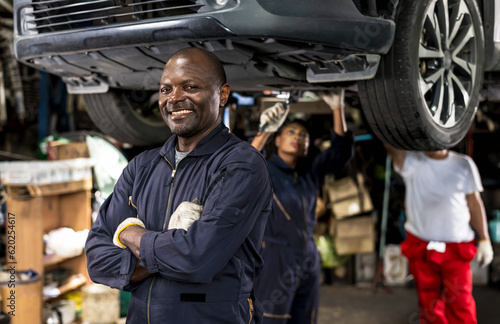 Happy black car mechanic standing with armfolded in auto repair with black mechanic woman working underneath car background, Car Mechanic
