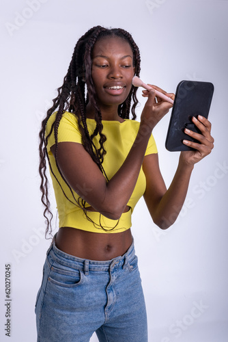 Mujer hermosa joven de color  que se maquilla delante de un pequeño espejo con un fondo blanco. Chica sonriente maquillándose el rostro antes de salir. 
