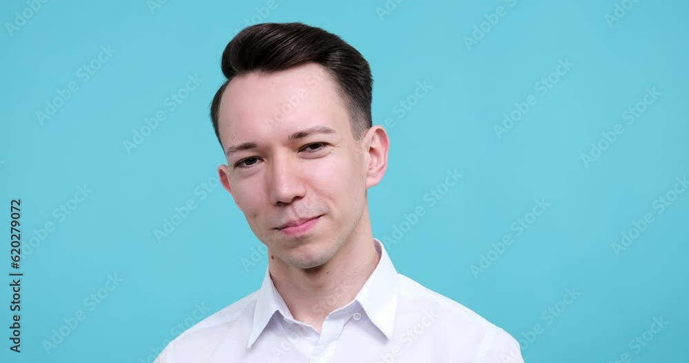 Against a blue background, a happy Caucasian man stands, his eyes locked on the camera. With a mischievous smile, he raises his index finger to his lips, making a playful shhh gesture. Against a blue background, a happy Caucasian man stands, his eyes locked on the camera. With a mischievous smile, he raises his index finger to his lips, making a playful shhh gesture.