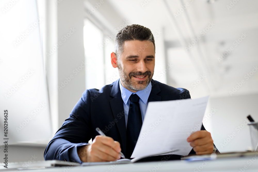 Positive middle aged businessman working with papers in office, reading financial reports and taking notes Positive middle aged businessman working with papers in office, reading financial reports and taking notes