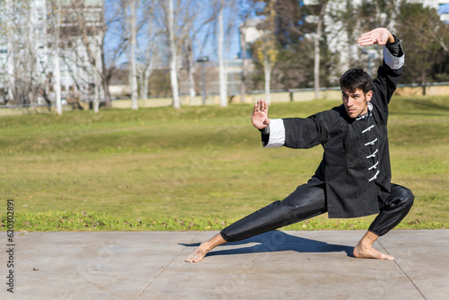 Young athletic martial arts fighter practicing kicks in a public park wearing classic kung fu uniform. Copy space