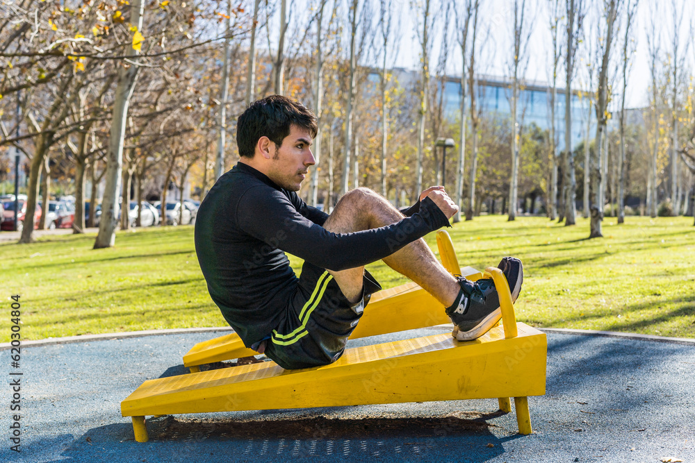 Confident young man doing abdominal exercises on a public equipment in outdoor fitness