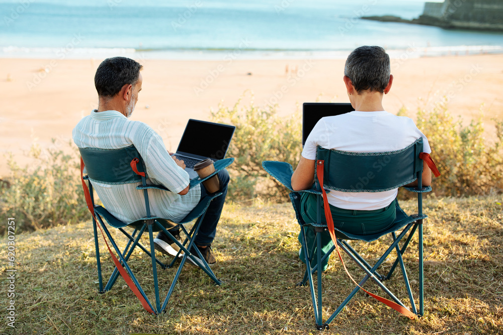 Mature Freelancers Couple Using Laptops At Ocean Coastline, Back View
