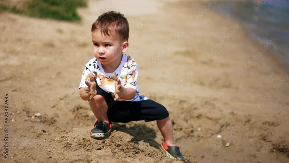 Baby boy playing with sand at the seashore. Kid is unhappy to have sand on his hands and claps them. Baby boy playing with sand at the seashore. Kid is unhappy to have sand on his hands and claps them.