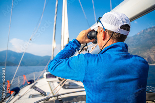 Young man captain on the yacht looking through binoculars during sailing boat control. Travel and active life.