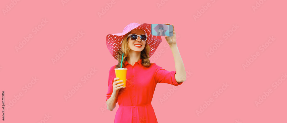 Summer portrait of beautiful happy smiling young woman taking selfie with smartphone and cup of juice wearing straw hat, pink dress on background