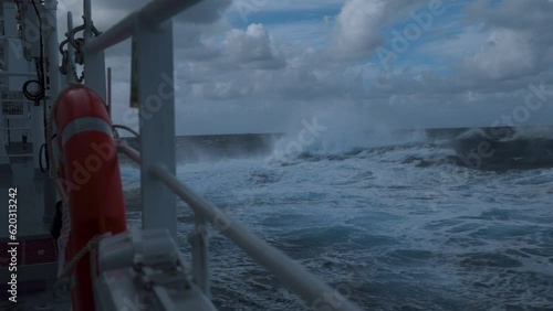 High waves and splashes along side. Ship in storm. White foam on sea. Deck railing and lifebuoy. Strong pitching. Vessel climb up wave.