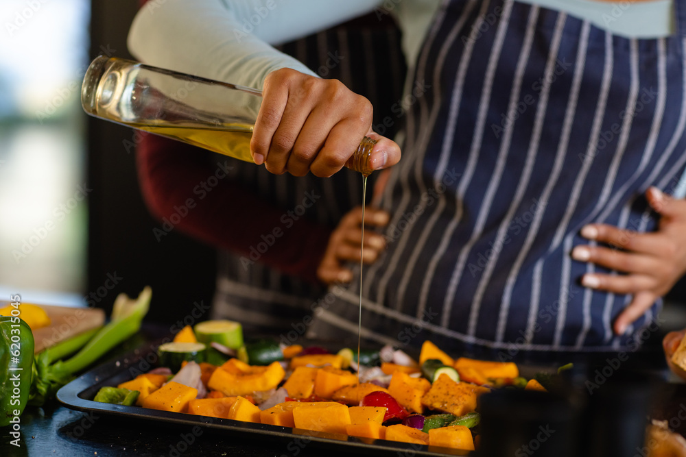 Mid section of biracial lesbian couple embracing and pouring oil on vegetables in kitchen Mid section of biracial lesbian couple embracing and pouring oil on vegetables in kitchen