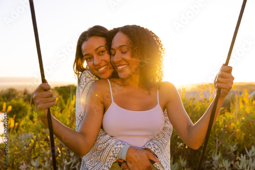 Happy biracial lesbian couple hugging and swinging on swing in sunny garden