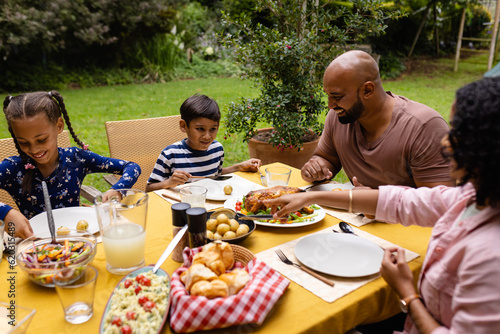 Happy biracial parents, son and daughter sitting at table having meal in garden