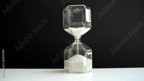 Hourglass on a white table and black background. Faceted modern hourglass with light gray sand. Hourglass on a white table and gray background. Faceted modern hourglass with light gray sand.
