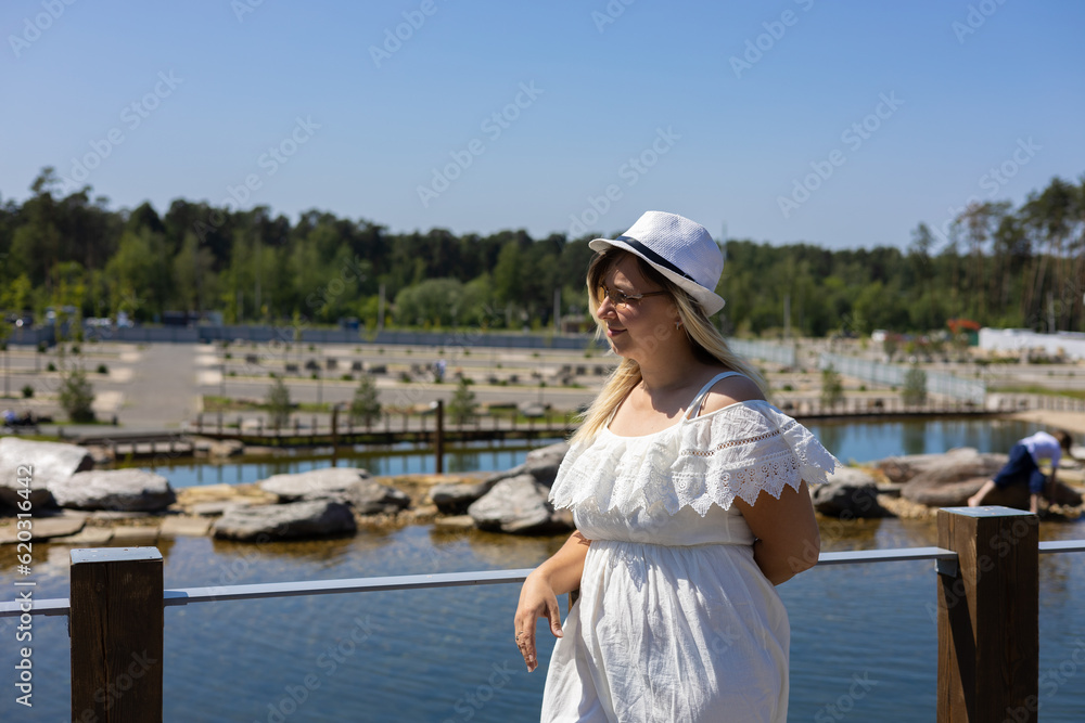 A blonde in a white dress on a pier against the background of water on a clear summer day. A woman travels through the park in summer. A woman stands on a bridge near a lake in a summer park.