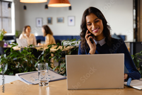 Happy middle eastern casual businesswoman using laptop and talking on smartphone at desk in office