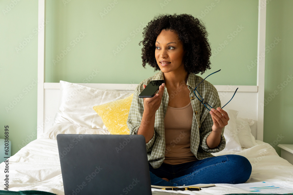 Biracial woman doing paperwork using laptop and talking on smartphone on bed in bedroom Biracial woman doing paperwork using laptop and talking on smartphone on bed in bedroom