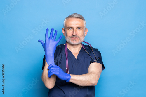 Doctor in medical uniform putting on protective sterile blue gloves while standing against blue background.