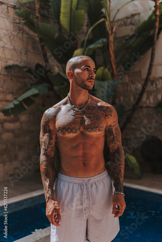 Chico joven tatuado y musculoso posando con bañador urbano en piscina de hotel un día soleado