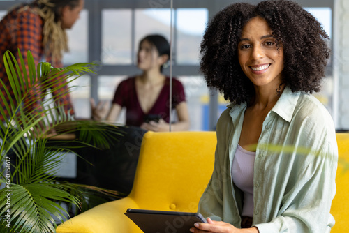 Portrait of happy biracial casual businesswoman on sofa, holding tablet and smiling at office