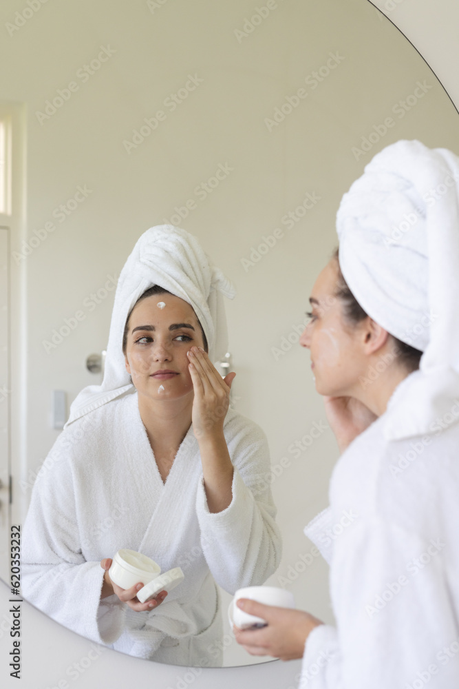 Happy caucasian woman with towel on head applying cream on face in bathroom Happy caucasian woman with towel on head applying cream on face in bathroom