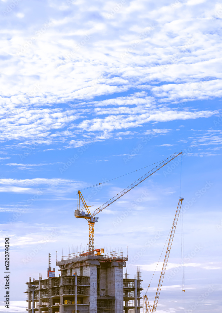 Tower cranes are working on top of high building structure against white clouds on blue sky in minimal style and vertical frame Tower cranes are working on top of high building structure against white clouds on blue sky in minimal style and vertical frame
