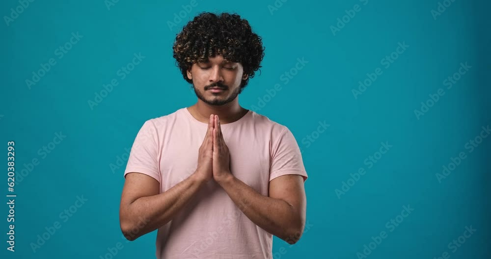 Young Indian man praying with eyes closed standing isolated on blue studio background Young Indian man praying with eyes closed standing isolated on blue studio background