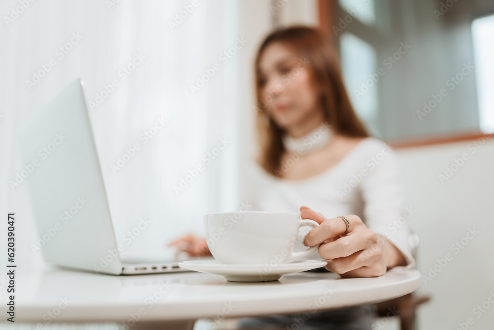 Selective focus hand picking up coffee cup, Businesswoman working online with computer laptop at coffee shop. Asian female relaxing with coffee in the morning. Selective focus hand picking up coffee cup, Businesswoman working online with computer laptop at coffee shop. Asian female relaxing with coffee in the morning.