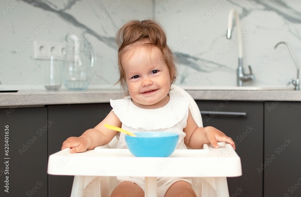 Baby is trying to eat on her own from plate, sitting on high chair. Hand with spoon for vegetable lunch. Weaning baby, first solid food for small child. Healthy baby food Baby is trying to eat on her own from plate, sitting on high chair. Hand with spoon for vegetable lunch. Weaning baby, first solid food for small child. Healthy baby food