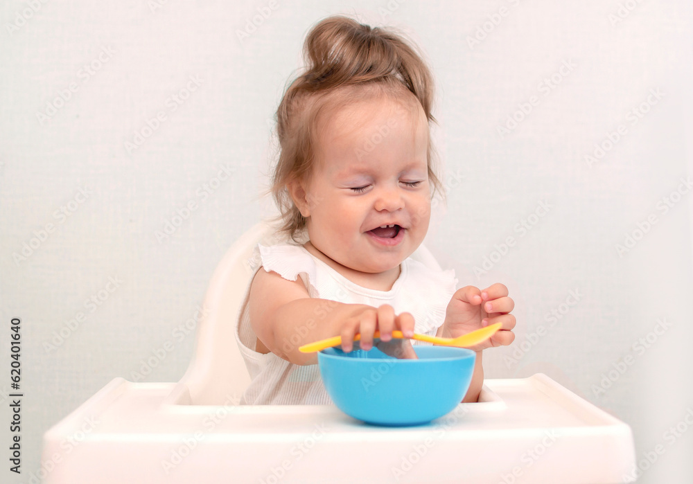 Baby is trying to eat on her own from plate, sitting on high chair. Hand with spoon for vegetable lunch. Weaning baby, first solid food for small child. Healthy baby food Baby is trying to eat on her own from plate, sitting on high chair. Hand with spoon for vegetable lunch. Weaning baby, first solid food for small child. Healthy baby food