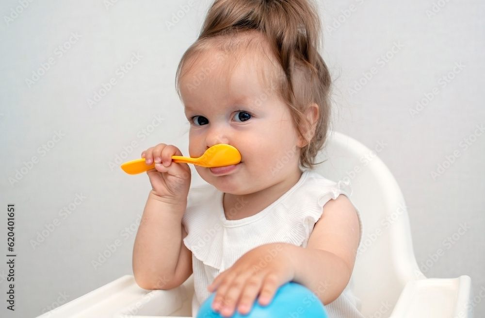 Baby is trying to eat on her own from plate, sitting on high chair. Hand with spoon for vegetable lunch. Weaning baby, first solid food for small child. Healthy baby food Baby is trying to eat on her own from plate, sitting on high chair. Hand with spoon for vegetable lunch. Weaning baby, first solid food for small child. Healthy baby food